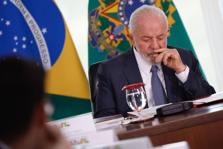 Sentado diante de uma mesa, com bandeira ao fundo, Lula, um homem branco, cabelo e barba da mesama cor. Ele veste um terno cinza, gravata e camisa branca e mexe na boca com a mão esquerda. Na mesa, diante dele, um copo com água e folhas de papel. 