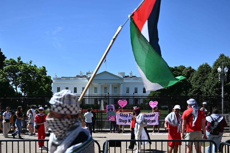Um grupo de manifestantes se reúne em frente à Casa Branca, com um indivíduo em primeiro plano segurando uma bandeira palestina. Cartazes com mensagens de protesto contra ações de Israel são visíveis, enquanto turistas e outros manifestantes se misturam ao fundo, sob um céu claro.