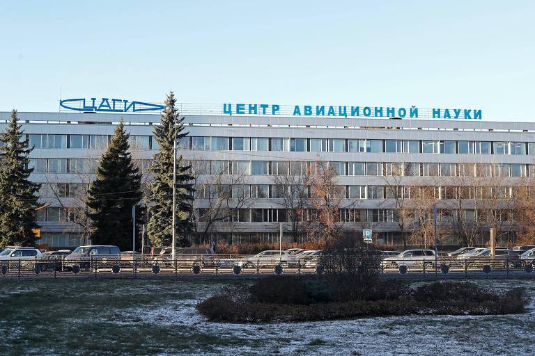 A fachada de um edifício moderno com letras na fachada, em russo, que se traduzem como "Centro de Ciência da Aviação", sob um céu claro. A arquitetura é complementada por árvores perenes e um pouco de neve no gramado