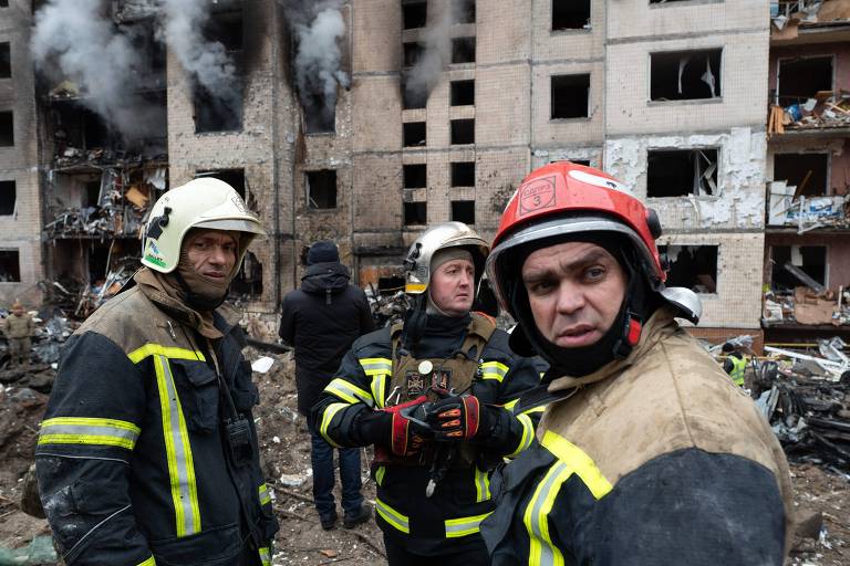 Três bombeiros -usando uniformes resistentes ao fogo,um deles, à direita da foto, com capacete vermelho, e outros dois com capacetes brancos- em frente a um prédio gravemente danificado, com fumaça ainda saindo das janelas quebradas