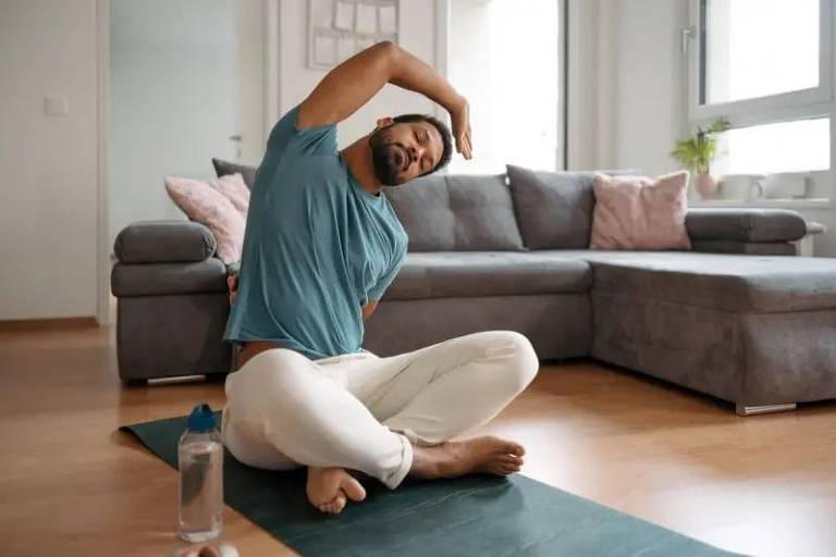 Um homem pratica yoga na sala de estar, executando um alongamento lateral enquanto está sentado sobre um tapete de yoga