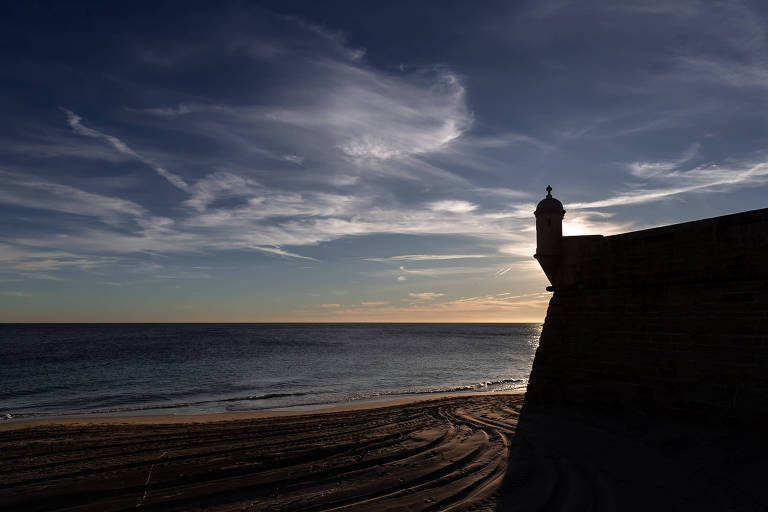 Imagem mostra final de tarde em praia deserta; grafia na areia é vista no contra luz de uma edificação à direita e o mar e céu azul à esquerda.