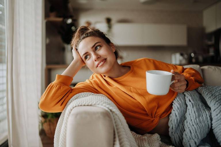 Uma mulher relaxa em um sofá aconchegante, segurando uma caneca branca e olhando para o lado. Ela está confortável em um moletom laranja, com uma manta de tricô azul sobre as pernas, sugerindo um momento de tranquilidade
