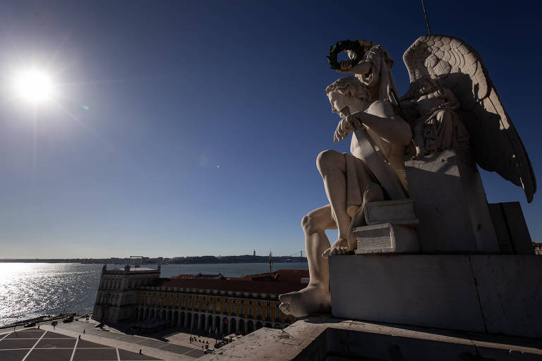 Imagem aérea mostra o rio Tejo do ponto de vista do alto de uma escultura, vista em primeiro plano. Céu azul e o sol em destaque.