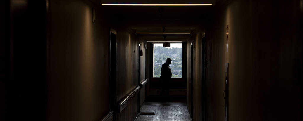 A imagem captura um momento introspectivo de uma pessoa parada ao final de um corredor escuro, olhando para fora de uma janela iluminada. A luz natural que entra pela janela contrasta com a penumbra do corredor, criando uma atmosfera de isolamento e reflexão.