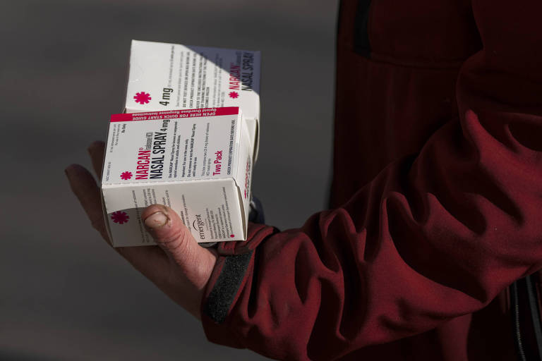 Uma pessoa com uma jaqueta vermelha segura firmemente várias caixas de medicamentos, onde se lê "Narcan" e "Emergency Medical Kit"