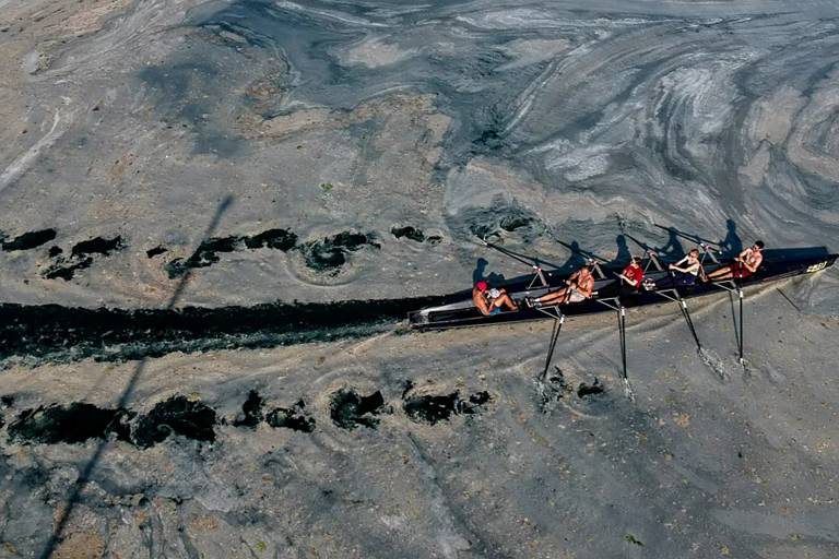 Imagem aérea de um caiaque com uma equipe de remo de cinco pessoas que desliza sobre água cinza, em um cenário que desafia a percepção, onde a água apresenta uma superfície cinzenta e texturizada