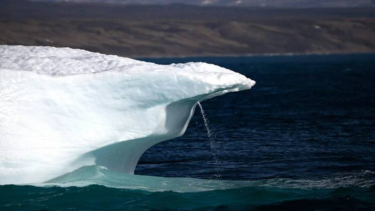 Um pedaço de iceberg flutua em águas azuis escuro, com uma pequena cascata de água formando uma esguicho em sua borda curva