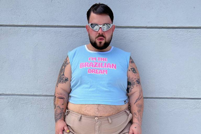 Um homem branco e gordo posa contra uma parede cinza. Ele veste uma regata azul claro com a frase 'I'm the Brazilian Dream' e uma bermuda cáqui, complementados por botas cano curto bege e óculos de sol espelhados. Seus braços são tatuados e as unhas pintadas de amarelo