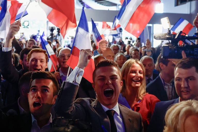 Um grupo de pessoas comemora em um evento, com expressões de alegria e entusiasmo, agitando bandeiras francesas. A atmosfera é de celebração e patriotismo, com um foco particular em um jovem homem no centro, que parece estar liderando o grupo em um momento de exaltação coletiva.