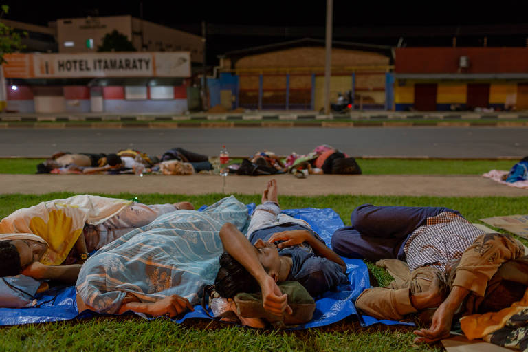 Fotografia mostra quatro refugiados deitados lado a lado em um gramado envolta de uma calçada. Estirados encima de lonas, alguns usam plásticos como cobertor, outros estão apenas com a roupa do corpo. Do outro lado da calçada, mais pessoas estão deitadas na mesma condição. Ao fundo, estabelecimentos da cidade. 