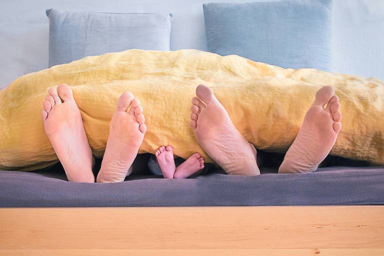 A imagem mostra os pés de dois adultos e um bebê emergindo de sob um cobertor amarelo no final da cama. A cama está arrumada com lençóis cinza e travesseiros combinando,
