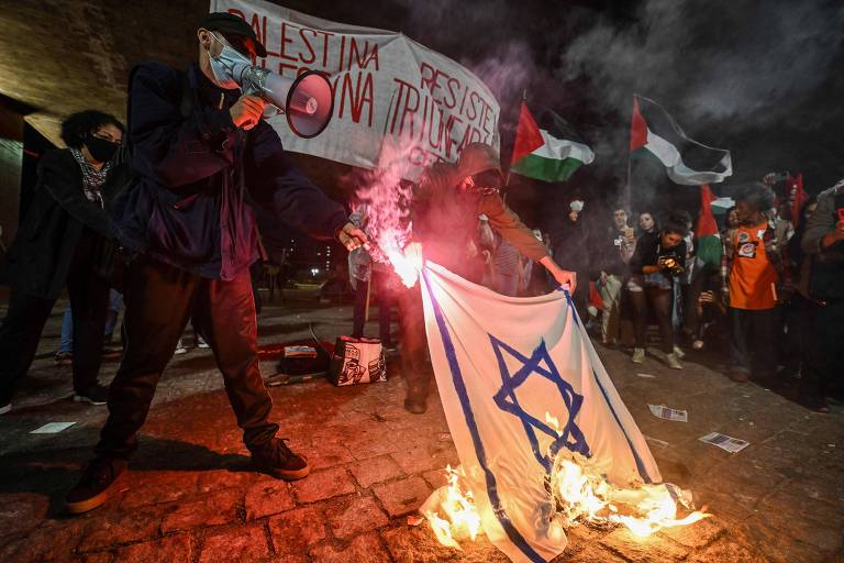 Manifestantes reunidos à noite em um protesto, onde uma bandeira de Israel está sendo queimada. Alguns participantes usam máscaras e seguram sinalizadores, enquanto outros agitam bandeiras da Palestina. O ambiente é carregado de tensão.