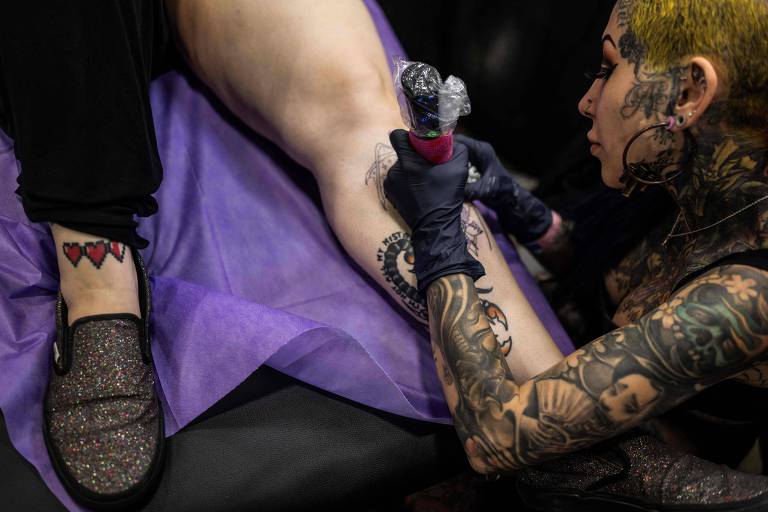 Um tatuador profissional, coberto por tatuagens da cabeça aos pés, está concentrado no processo de tatuar a perna de um cliente. O ambiente sugere um estúdio de tatuagem, com o cliente relaxado enquanto o artista trabalha com precisão, usando luvas pretas e segurando uma máquina de tatuagem.
