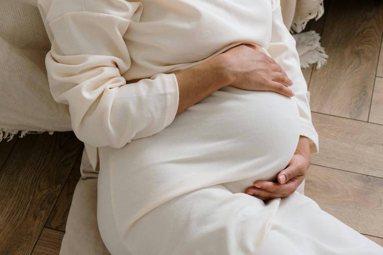 A imagem mostra uma mulher grávida sentada, com as mãos apoiadas sobre a barriga. Ela está vestindo uma roupa clara e confortável, e o ambiente ao redor parece ser acolhedor, com piso de madeira e um tecido claro ao fundo.
