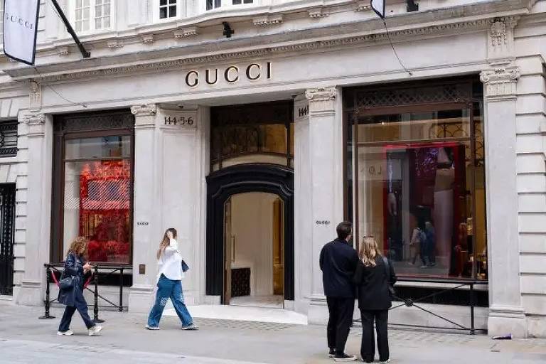 Transeuntes caminham diante de uma loja da Gucci, com sua fachada clássica e elegante. A vitrine exibe itens de luxo, enquanto um casal parece contemplar a entrada