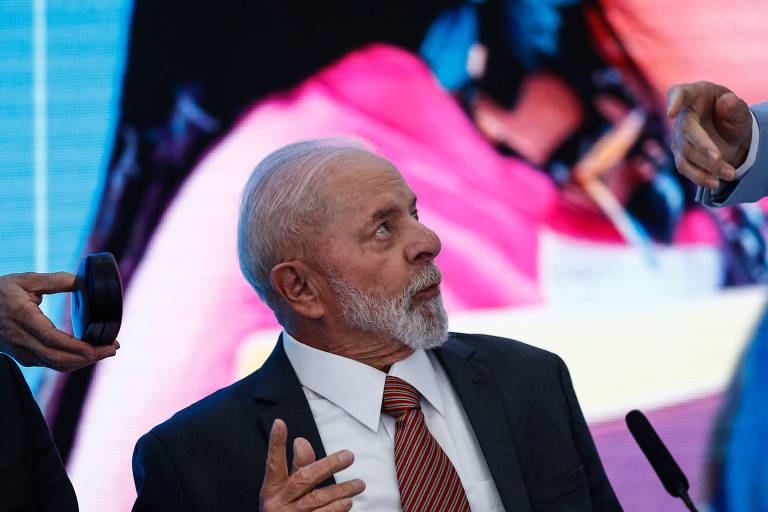 Lula, um homem com barba branca e cabelos grisalhos, vestindo terno escuro com gravata vermelha, gesticula enquanto fala, com uma expressão séria e concentrada. Ao fundo, um telão exibe imagens desfocadas com predominância de cores azul e rosa