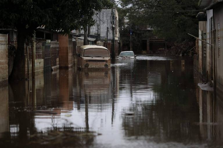 A imagem mostra uma rua residencial inundada, com águas paradas refletindo as casas e árvores ao redor. Dois veículos estão parcialmente submersos, evidenciando a gravidade da inundação