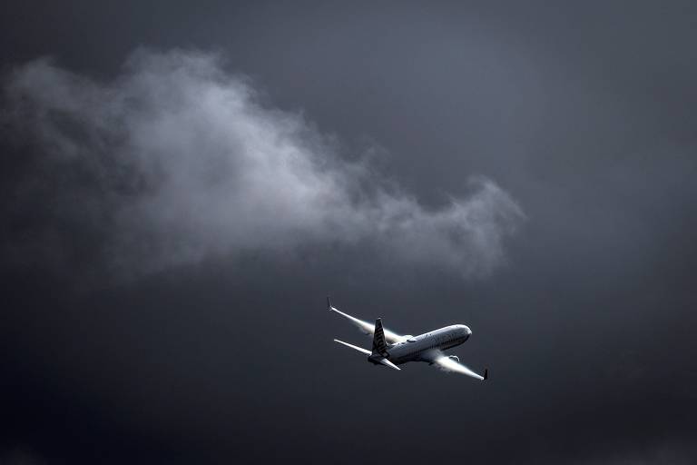 Um avião comercial emerge sob um céu nublado e sombrio, destacando-se como um ponto brilhante contra o fundo escuro.