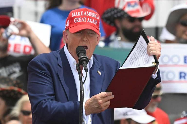 Trump está discursando em um evento ao ar livre, vestindo um boné vermelho com o slogan "Make America Great Again". Ele segura um caderno. Ao fundo, pode-se ver uma multidão de apoiadores, alguns usando bonés semelhantes e segurando cartazes de apoio.