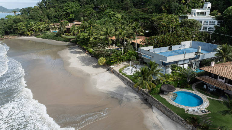 Uma vista aérea revela um trecho de praia, onde a floresta tropical encontra o oceano. As ondas surgem nas margens da areia. A imagem mostra luxuosas residências e uma piscina azul-turquesa e o verde da vegetação circundante.