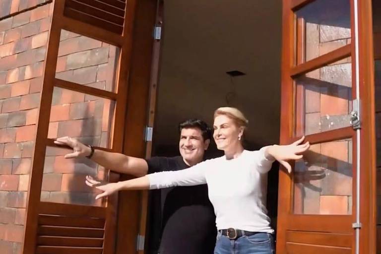Um homem e uma mulher abrem uma janela juntos. O homem veste uma camiseta preta enquanto a mulher uma blusa de manga longa branca