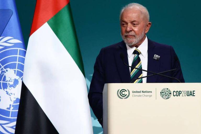 Um homem com expressão séria discursa em um púlpito com os logotipos da ONU e da COP28, com a bandeira dos Emirados Árabes Unidos ao fundo.