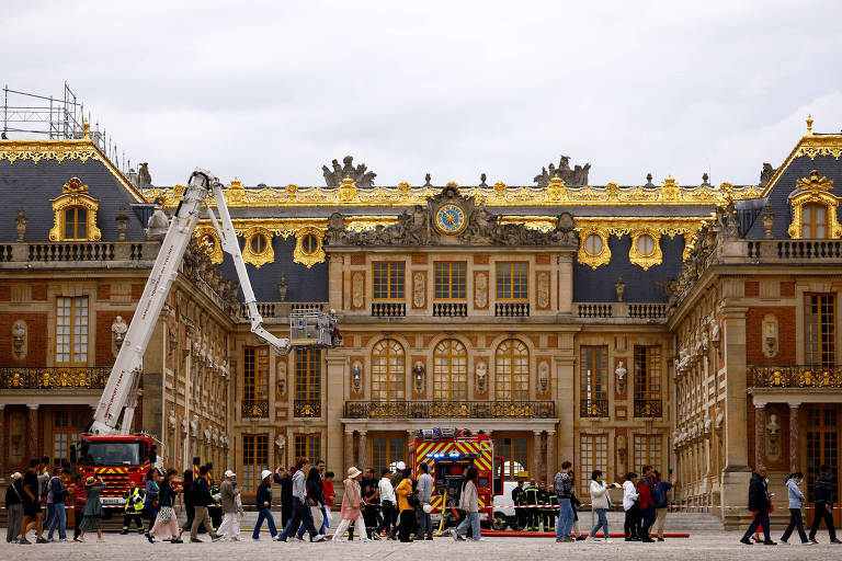 Uma cena diante do histórico Palácio de Versalhes mostra a combinação do cotidiano e da conservação patrimonial, com turistas passeando enquanto uma equipe realiza trabalhos de manutenção utilizando uma escada de bombeiro.