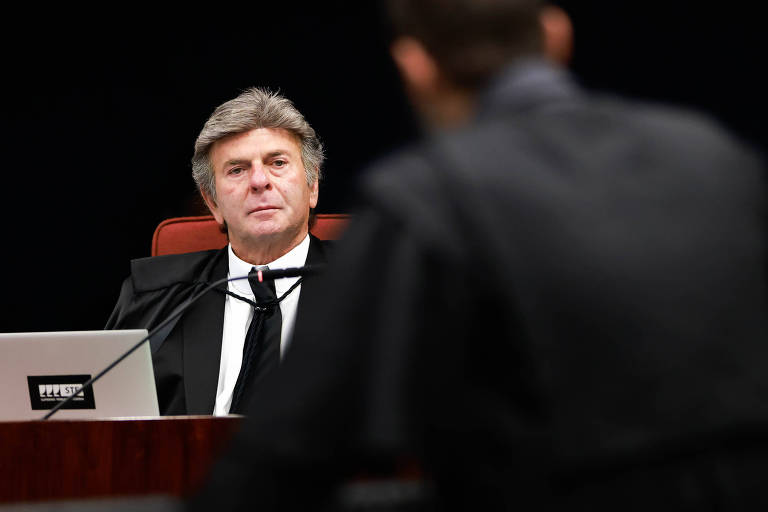 Fotografia mostra o ministro Luiz Fux durante sessão na Primeira Turma do STF. Ele usa terno preto, camisa social branca e gravata preta, e aparece olhando para outro homem, que está de costas e com a aparência desfocada.