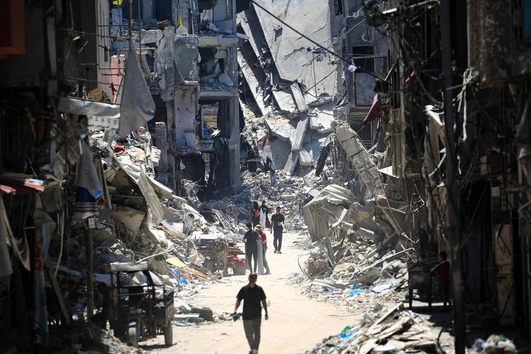 Palestinos caminham em rua com edifícios destruídos na cidade de Khan Yunis, na Faixa de Gaza