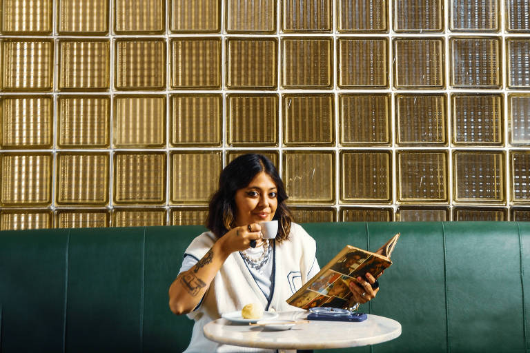 Uma pessoa está sentada em uma mesa de café, imersa na leitura de um livro. A iluminação suave e a parede de azulejos geométricos criam um ambiente acolhedor e estiloso, enquanto um círculo luminoso acima adiciona um toque ao cenário.
