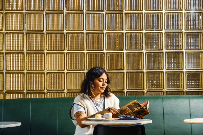 Uma mulher está sentada em um banco de estofado verde enquanto lê um livro. Ela está em um ambiente com paredes adornadas por blocos de vidro translúcido e dourado. Uma xícara de café e um prato com um pedaço de bolo a acompanham.