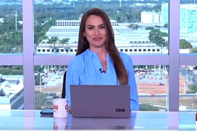  Uma apresentadora de notícias sorridente, vestida com uma camisa azul, em frente a um laptop e canecas com o logotipo da CNN, com uma vista panorâmica da cidade ao fundo através de uma janela ampla.