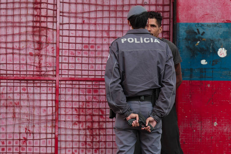 Um policial de costas, vestido com uniforme cinza em um interrogatório com um civil contra uma parede. Nas costas o policial segura um revólver com as duas mãos.