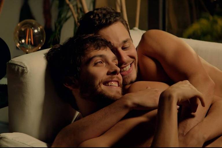 Dois homens aparecem abraçados em um sofá. Eles estão sorrindo e parecem estar desfrutando da companhia um do outro, em um momento de relaxamento e intimidade.