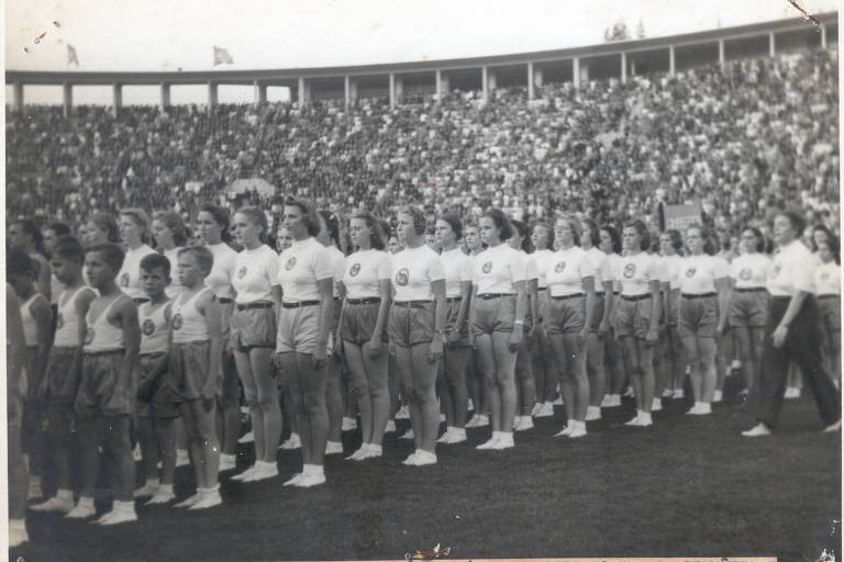 A imagem em preto e branco captura um momento solene durante a inauguração do Estádio do Pacaembu em 1940, mostrando equipes do Sport Club Germânia alinhadas no campo, vestindo uniformes de ginástica, sob o olhar atento de uma multidão que preenche as arquibancadas ao fundo.