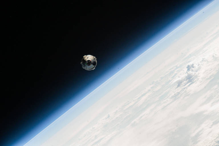 Uma cápsula flutua no espaço, com a curvatura da Terra e a fina camada da atmosfera delineando o horizonte abaixo.
