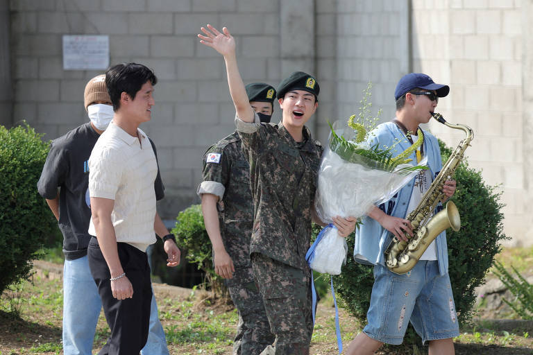 Imagem mostra homem de uniforme militar segura um bouquê de flores e acena com a mão. Andando ao seu lado, quatro homens. Um deles toca saxofone.