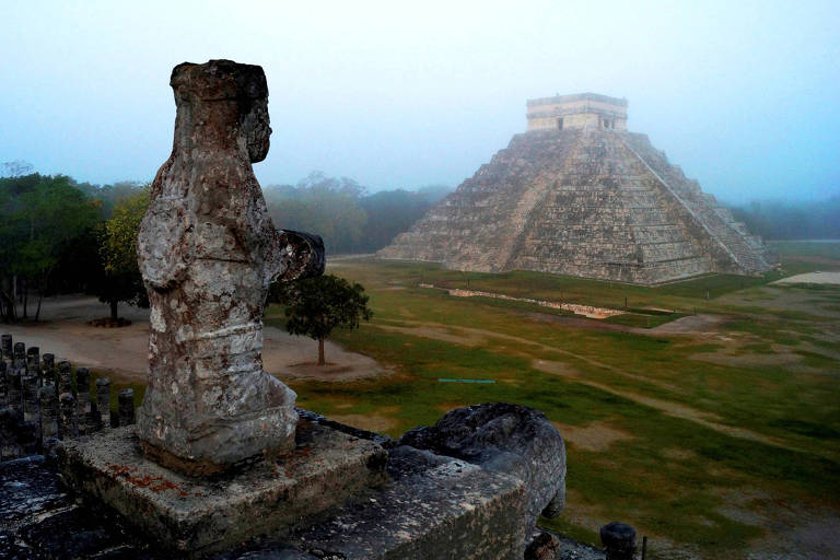 Uma estátua de pedra desgastada pelo tempo em primeiro plano, com a pirâmide de Kukulkán ao fundo, envolta pela suave névoa do amanhecer em Chichén Itzá, um dos mais famosos sítios arqueológicos da civilização maia.
