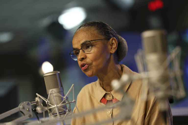 Uma mulher com expressão concentrada, usando blusa bege, óculos e cabelos grisalhos presos em um coque, fala ao microfone em um estúdio de rádio, cercada por equipamentos de transmissão. 