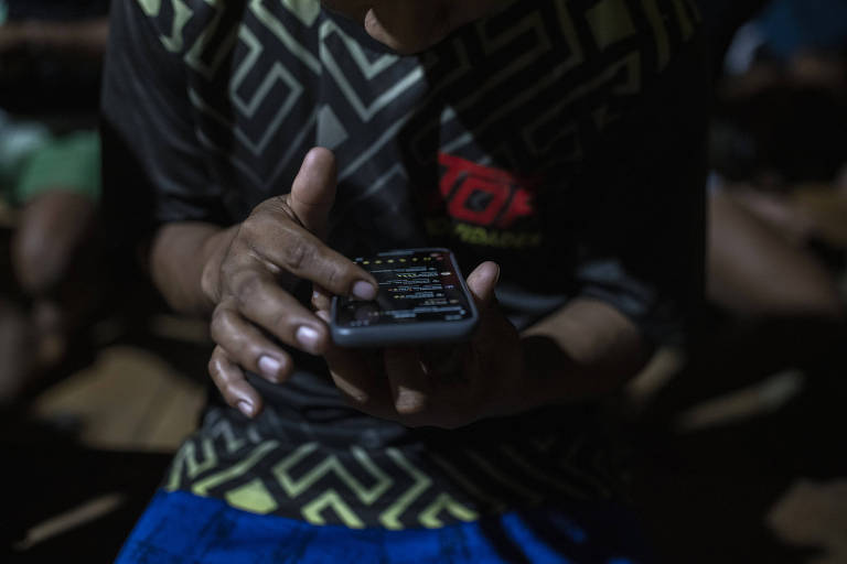 A imagem mostra as mãos de uma pessoa segurando um smartphone com a tela acesa, em um ambiente escuro. A luz do dispositivo ilumina parcialmente as mãos e a roupa da pessoa, criando um contraste com o fundo