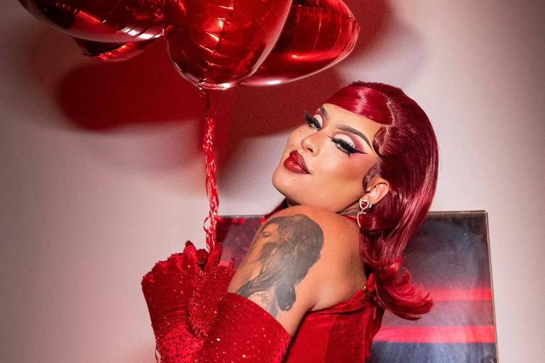 Uma mulher exibe um visual em tons de vermelho, com um conjunto de balões em forma de coração flutuando ao seu lado. Seu cabelo vermelho combina com a maquiagem marcante e o vestido , enquanto ela segura a fita dos balões.