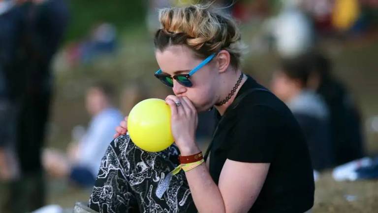 Uma mulher com óculos escuros e cabelo preso em um coque alto aspira o gás de um balão amarelo em um ambiente ao ar livre