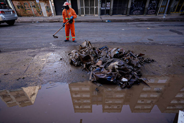 Na foto, um gari de roupa laranja dos pés à cabeça com uma varroura na mão está próximo de uma pilha de entulho, acumulada em uma rua parcialmente alagada