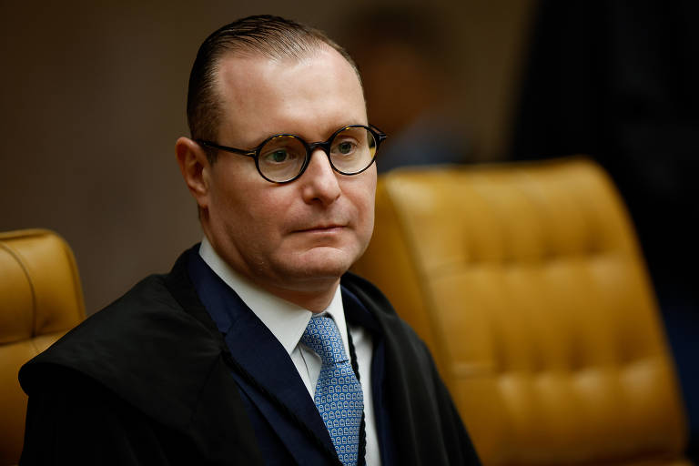 Um homem com cabelo curto e liso, usando óculos de armação preta, está sentado em uma cadeira de tribunal. Ele veste um terno escuro com uma gravata clara e um manto judicial. O fundo apresenta uma cadeira de couro marrom e um ambiente de tribunal.