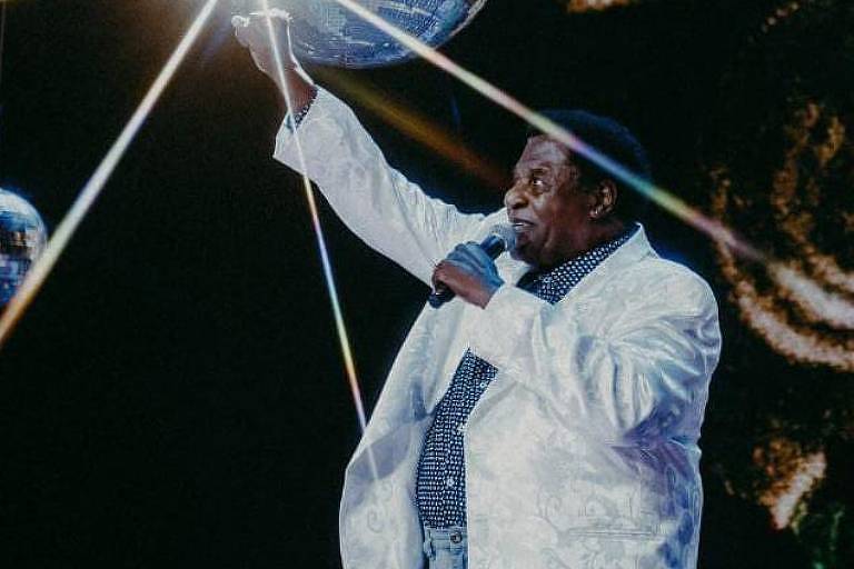 A imagem mostra um cantor negro e vestido com uma roupa branca, com microfone na mão em um palco