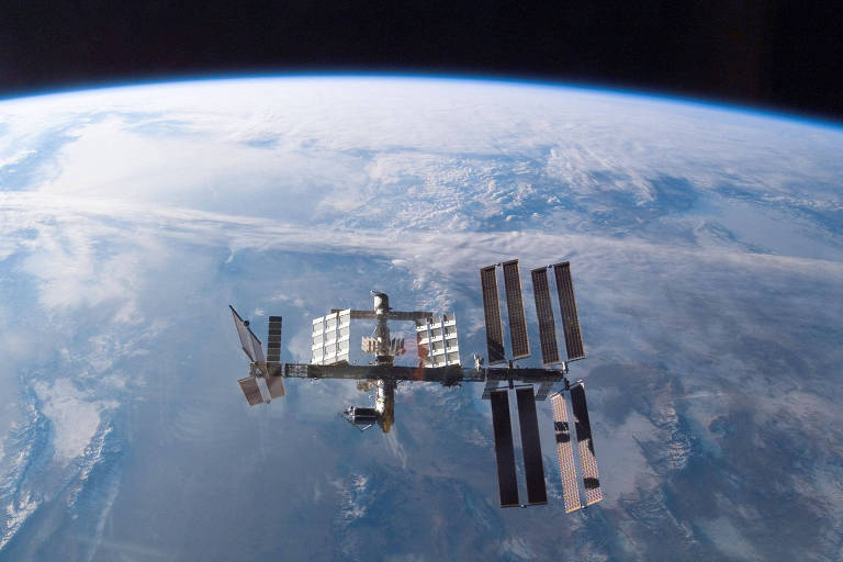 A imagem captura a Estação Espacial Internacional (ISS) em órbita acima da Terra, com o azul do oceano e as nuances da atmosfera terrestre ao fundo.
