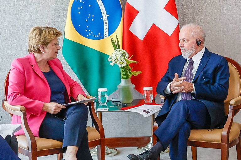 O presidente Lula e a presidente da Suíça estão sentados em cadeiras opostas em uma sala iluminada, envolvidos em uma conversa séria. Entre eles, uma mesa baixa suporta um arranjo de flores, e atrás de cada um, as bandeiras do Brasil e da Suíça estão posicionadas, simbolizando uma reunião bilateral entre as duas nações