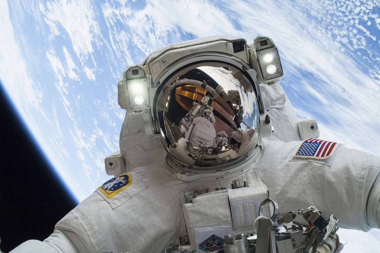 Um astronauta capturado em close-up flutua no vácuo do espaço, com a Terra azul e branca ao fundo. O reflexo no visor do capacete revela um vislumbre da estação espacial

