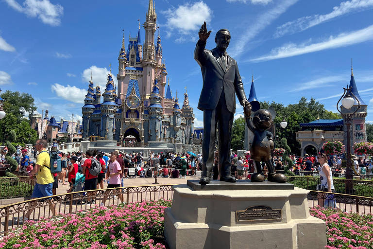 A imagem mostra uma estátua de bronze de Walt Disney segurando a mão de Mickey Mouse em primeiro plano, com o Castelo da Disney ao fundo. O castelo é decorado com torres azuis e douradas, e há muitas pessoas ao redor, algumas usando camisetas coloridas. O céu está claro com algumas nuvens brancas e há flores rosas na frente da estátua.
